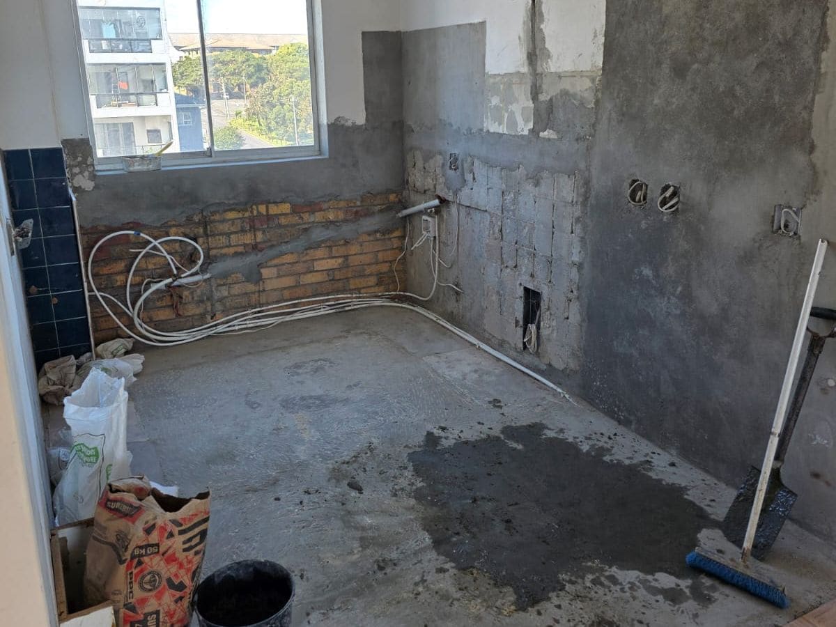 A stripped apartment kitchen with bare brick and plaster walls, plumbing runs along the floor and a window, before any cabinetry.