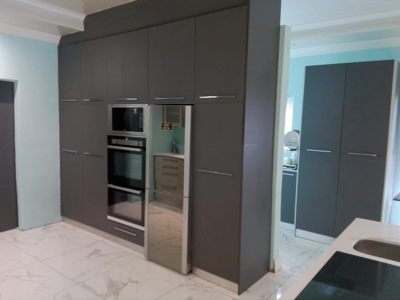 A wall of charcoal floor-to-ceiling units housing a built-in double oven, beside a pale blue wall, with the corner of a white island and sink in the foreground.